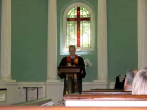A man in black robe standing at podium with cross on wall.